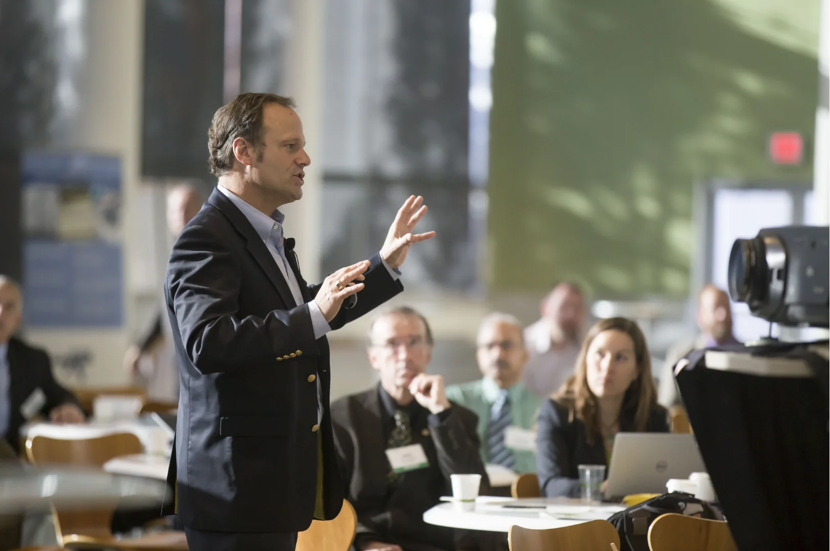A speaker mid-gesture, talking straight to camera in front of a small corporate audience