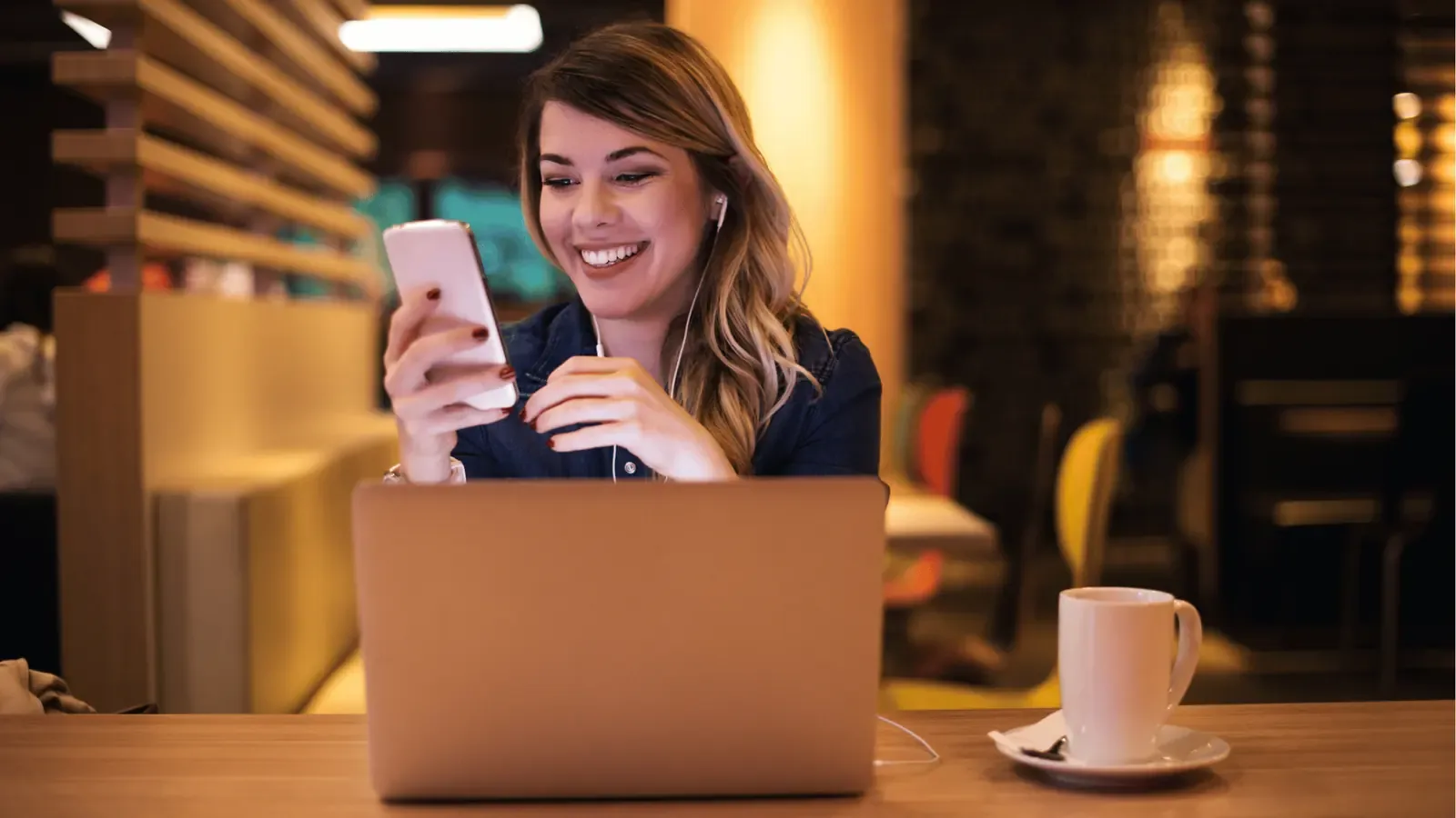 A woman smiling at her phone beside an open laptop in a cafe